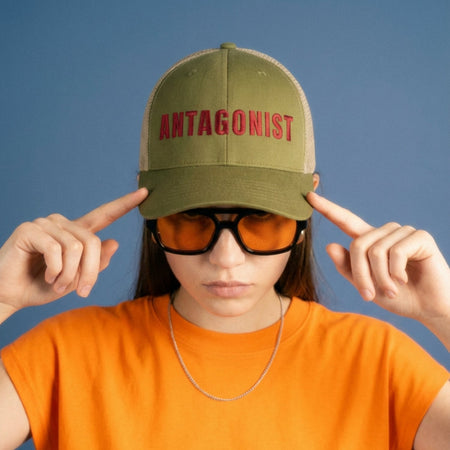 Person wearing an olive green cap with 'ANTAGONIST' text, orange shirt, and sunglasses against a blue background