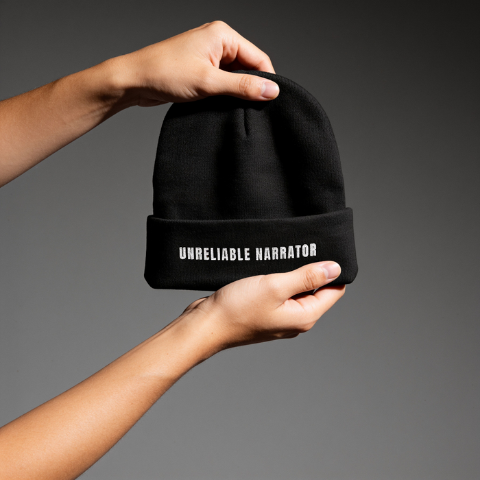 A person's hands holding a black knit beanie with the text 'UNRELIABLE NARRATOR' printed in white on the folded cuff, set against a dark gray gradient background.
