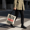 A person walking across a street crosswalk carrying a canvas tote bag with the text 'READ BANNED BOOKS' in black and red lettering. The person is wearing an olive green button-up shirt, black pants, and black leather boots.