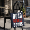 A navy blue canvas tote bag with black handles, featuring the text 'READ BANNED BOOKS' in white and distressed red lettering, hanging from a black metal chair in an outdoor cafe setting.