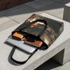 A tote bag with a dark classical painting print sits on a concrete bench outdoors, styled with a silver laptop, notebook, phone, and wallet.