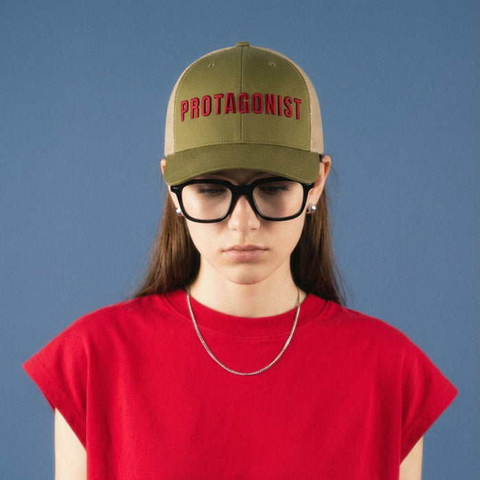 Person wearing a green cap with 'PROTAGONIST' text and a red crop top against a blue background