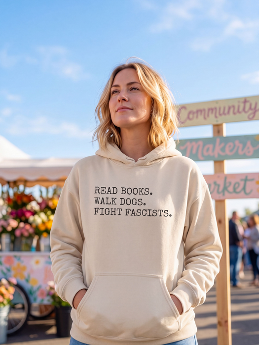 A woman wearing a cream-colored hoodie with the text 'READ BOOKS. WALK DOGS. FIGHT FASCISTS.