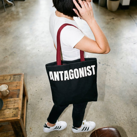 Person holding a black tote bag with 'ANTAGONIST' text in an indoor setting.