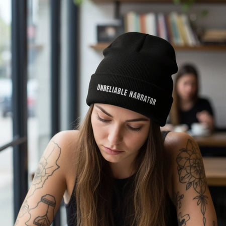Woman wearing a black beanie with 'Unreliable Narrator' text, writing in a notebook at a cafe.