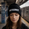Woman wearing a black beanie with 'ANTAGONIST' text on a subway platform.