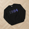 Black sweatshirt with '1984' printed on it, laid flat on a wooden floor.