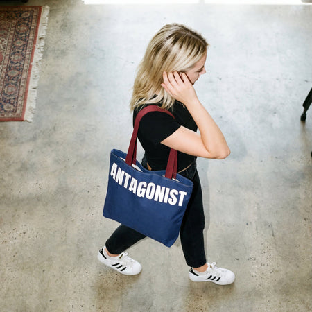 Woman holding a blue tote bag with 'ANTAGONIST' text in an indoor setting.