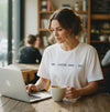  Woman in a white Screenwriter tee working on a laptop in a coffee shop with a cup of coffee.